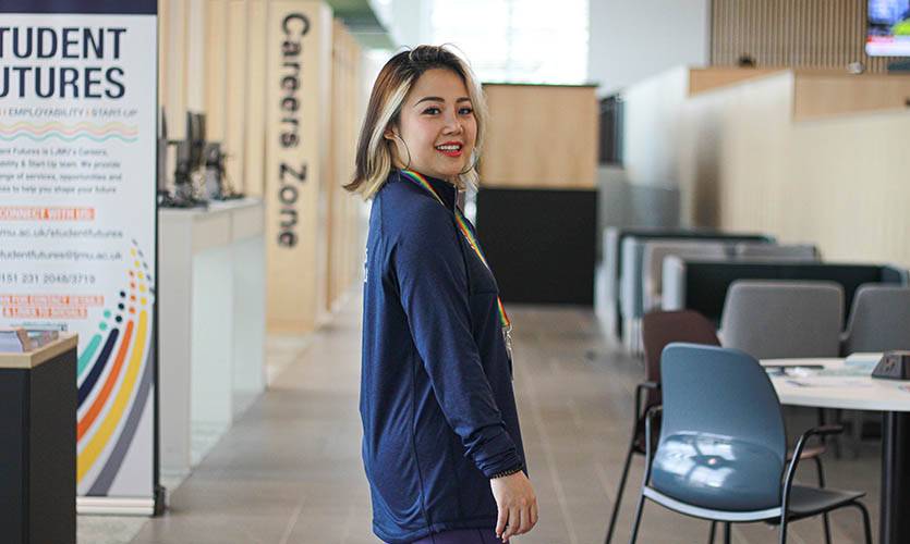 Gaby is pictured from the waist up wearing a navy-blue top and is stood side on looking at the camera and smiling, she has blonde highlights at the front of a cropped hair cut and is wearing red lipstick