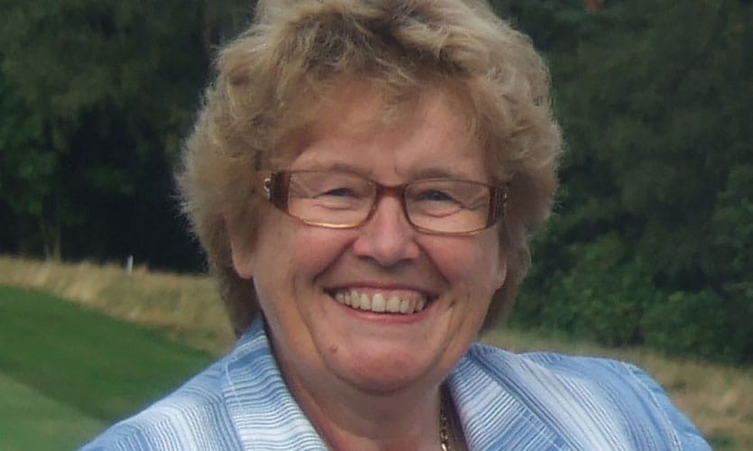 A head and shoulders photograph of Pat smiling as she looks to the camera wearing a pale blue t-shirt and short-sleeved jacket, she is also wearing metal framed glasses on her face