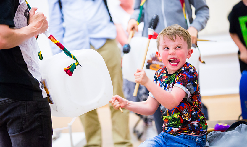 A small child grinning whilst drumming on a plastic container