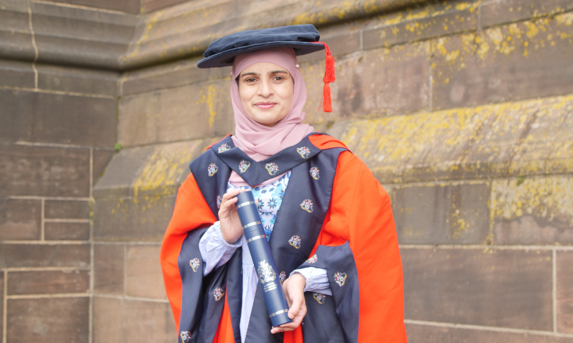 A woman wearing her graduation cap and gown and holding a scroll inbetween her hands